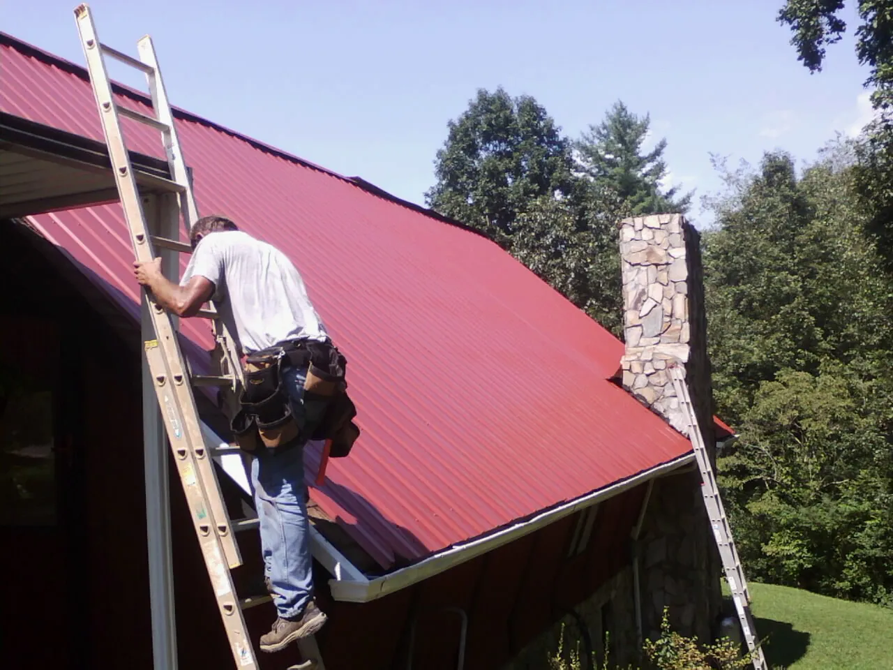 Roofer on a ladder beside a red metal roof and stone chimney — Soffit & Fascia Repair by Roofing Brothers in Stanfordville